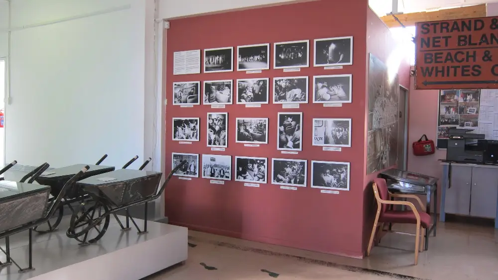 Plate 9. Wall with nineteen photos with labels and an artwork with wheelbarrows in front of it.
